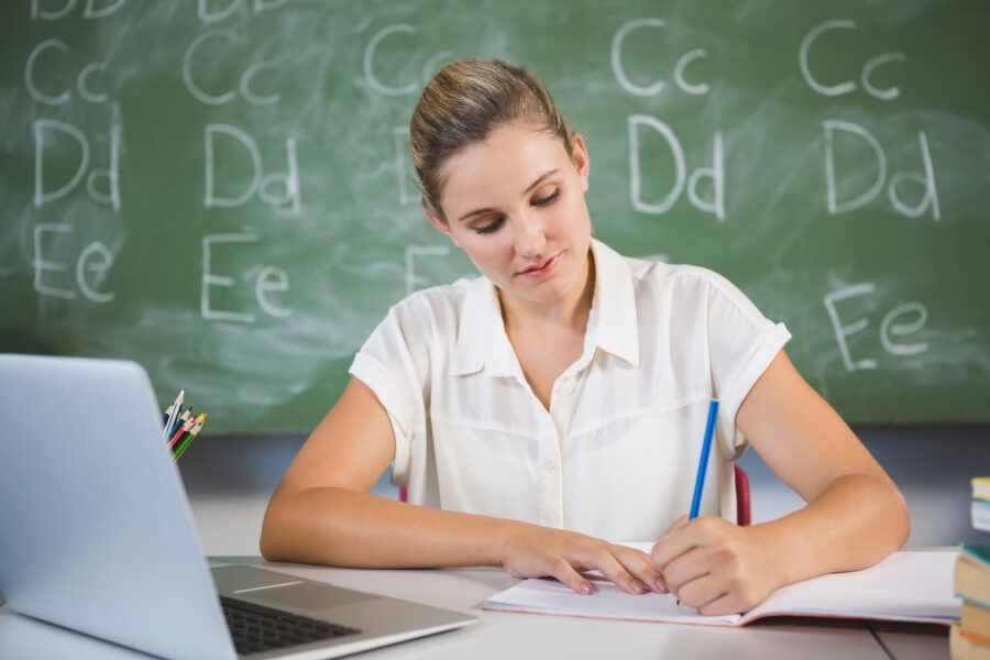 white teacher sitting at desk near computer in front of blackboard 9